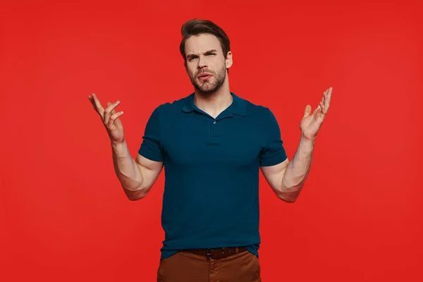 Jovem descontente em desgaste casual fazendo um rosto e gesto enquanto está de pé contra o fundo vermelho — Fotografia de Stock