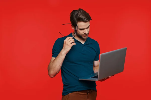 Bello giovane uomo in abbigliamento casual che trasporta laptop mentre in piedi sullo sfondo rosso — Foto Stock