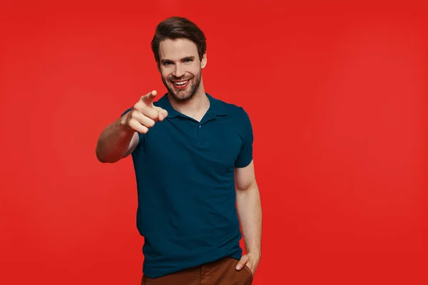 Happy young man in casual wear pointing camera while standing against red background — Stock Photo, Image