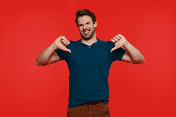 Displeased young man in casual wear looking at camera and showing his thumbs down — Stock Photo, Image