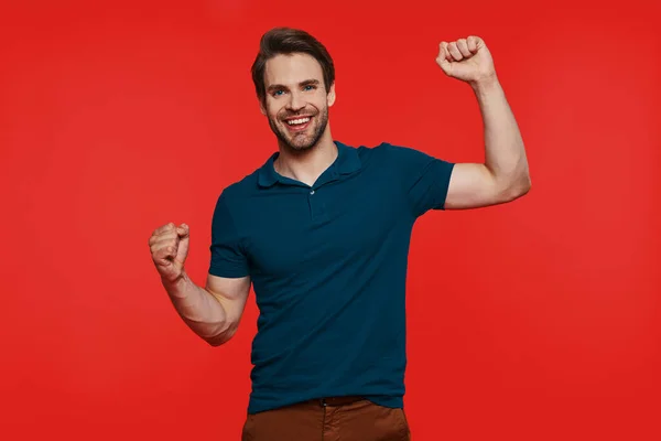 Happy young man in casual wear looking at camera and gesturing while standing against red background — Stock Photo, Image