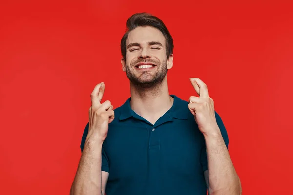 Glada ung man i casual wear hålla fingrarna korsade när du står mot röd bakgrund — Stockfoto