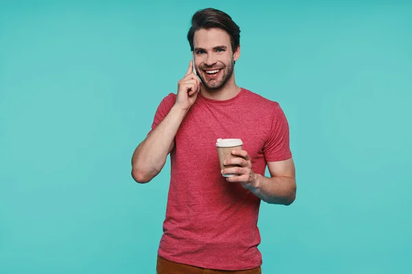 Bonito jovem sorrindo homem em roupas casuais segurando copo e falando no telefone inteligente — Fotografia de Stock