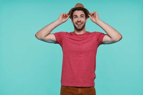 Handsome young man adjusting his fedora hat and smiling while standing against blue background — Stock Photo, Image