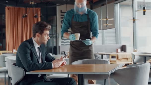 Camarero en protector facial trayendo al cliente una taza de café en el restaurante — Vídeo de stock