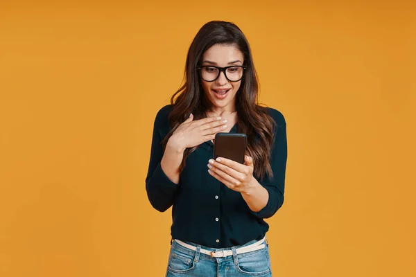 Mujer joven conmocionada con ropa casual usando un teléfono inteligente mientras está de pie contra el fondo amarillo — Foto de Stock