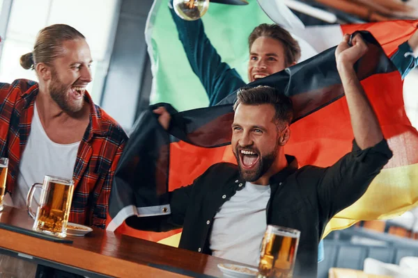 Junge Männer mit internationalen Fahnen genießen Bier, während sie in der Kneipe ein Sportspiel verfolgen — Stockfoto