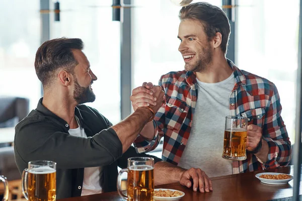 Dva mladí muži v neformálním oblečení potřásání rukou a usmívání se při vychutnávání piva u baru v hospodě — Stock fotografie
