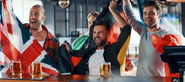 Vrolijke jonge mannen bedekt met internationale vlaggen genieten van bier tijdens het kijken naar sport wedstrijd in de pub — Stockfoto