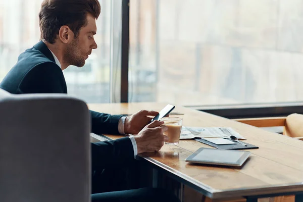 Vista lateral de joven hombre de negocios guapo en traje completo usando el teléfono inteligente y beber café — Foto de Stock