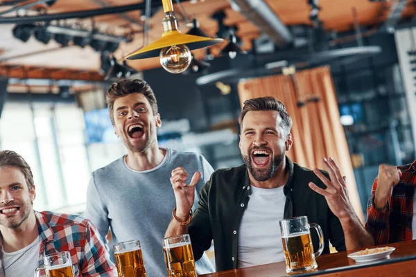 Animando a los jóvenes con ropa casual bebiendo cerveza y viendo el juego deportivo mientras están sentados en el pub — Foto de Stock