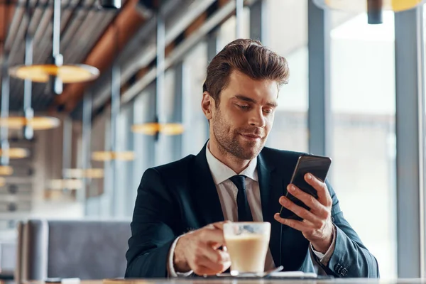 Fešák mladý muž v plném obleku pomocí chytrého telefonu a pití kávy při posezení v restauraci — Stock fotografie