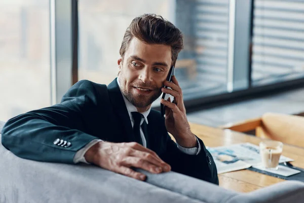 Guapo joven sonriente hombre en traje completo hablando en el teléfono inteligente y mirando por encima del hombro — Foto de Stock