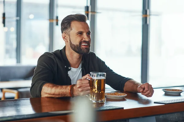 Jovem alegre em roupas casuais desfrutando de cerveja enquanto passa um tempo no pub — Fotografia de Stock