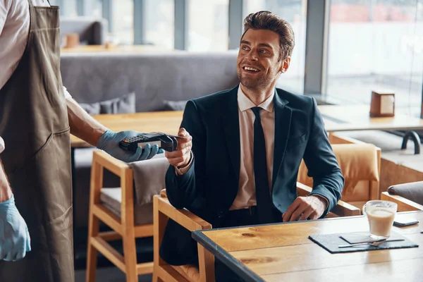 Przystojny młody uśmiechnięty mężczyzna w pełnym garniturze dokonujący płatności bezdotykowej siedząc w restauracji — Zdjęcie stockowe