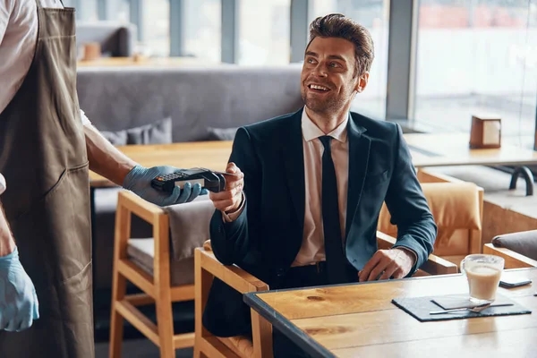 Handsome young smiling man in full suit making a contactless payment while sitting in the restaurant — 图库照片