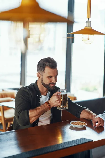 Schöner junger Mann in lässiger Kleidung genießt Bier und lächelt, während er Zeit in der Kneipe verbringt — Stockfoto