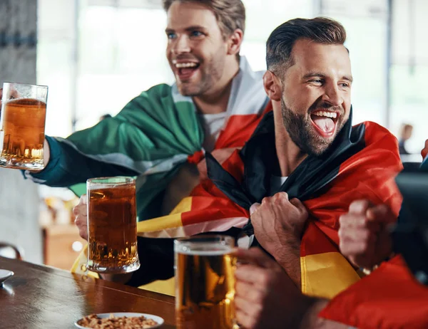 Jovens felizes cobertos de bandeiras internacionais brindam com cerveja enquanto assistem a jogos esportivos no pub — Fotografia de Stock