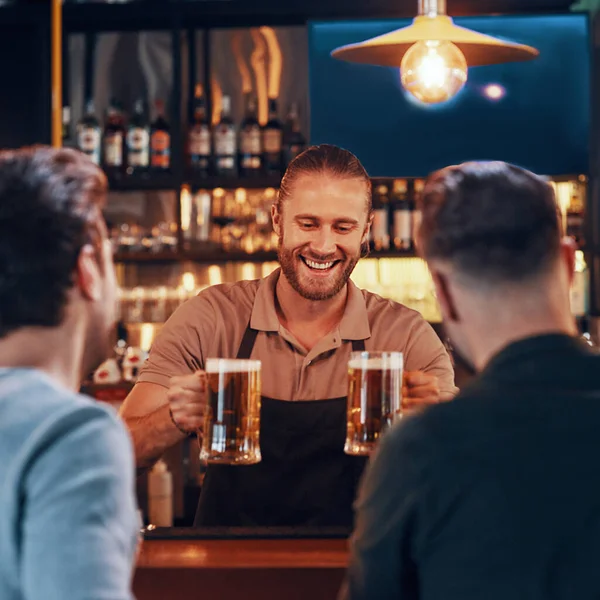 Fröhlicher Barkeeper, der jungen Männern Bier serviert, während er in der Kneipe an der Theke steht — Stockfoto