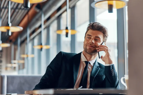 Bonito jovem em terno completo falando no telefone celular e sorrindo enquanto sentado no restaurante — Fotografia de Stock