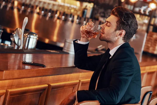 Schöner junger Mann in vollem Anzug trinkt Alkohol, während er an der Theke im Restaurant sitzt — Stockfoto