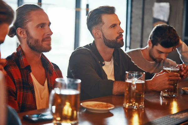 Grupo de jóvenes con ropa casual disfrutando de la cerveza mientras ven la televisión en el pub — Foto de Stock