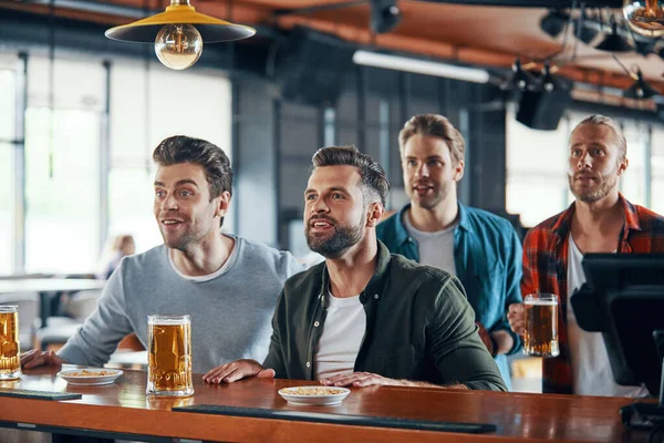 Аплодирующие молодые люди в повседневной одежде смотрят спортивную игру и наслаждаются пивом, сидя в пабе — стоковое фото