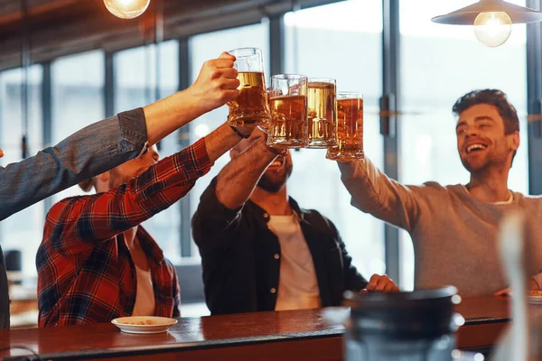 Šťastní mladí muži v neformálním oblečení připíjeli jeden na druhého pivem a usmívali se — Stock fotografie