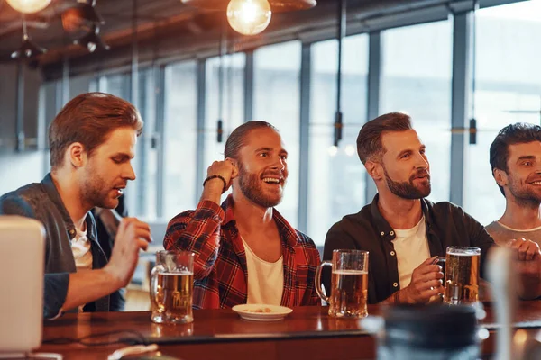 Grupo de jovens felizes em roupas casuais desfrutando de cerveja enquanto sentados no balcão do bar no pub — Fotografia de Stock