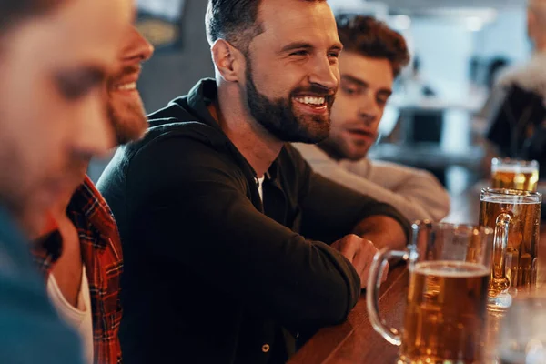 Gelukkig jonge mannen in casual kleding genieten van bier en glimlachen terwijl zitten in de in de pub samen — Stockfoto