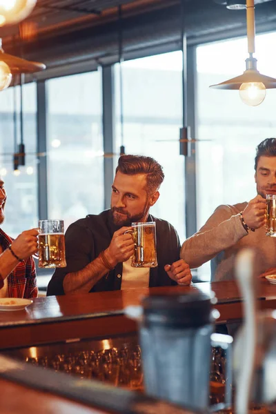 Guapos hombres jóvenes con ropa casual disfrutando de la cerveza mientras están sentados en el mostrador del bar en el pub —  Fotos de Stock