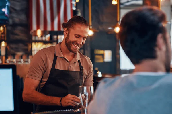 Young male bartender cleaning glasses and smiling while standing at the bar counter in pub — 图库照片