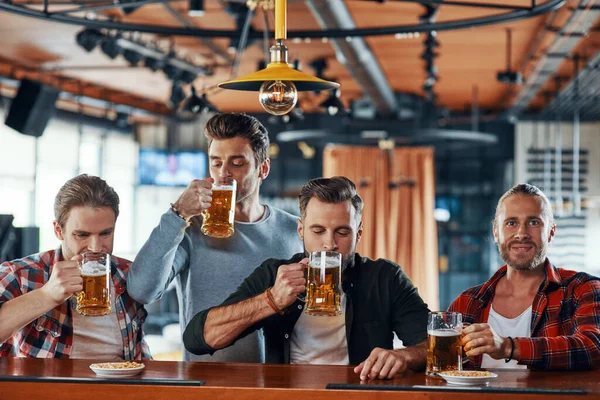 Group of cheerful young men in casual clothing enjoying beer while sitting at the bar counter in pub — 图库照片