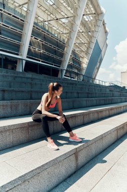 Spor kıyafetli güzel bir kadın dışarıda oturuyor ve başka tarafa bakıyor.