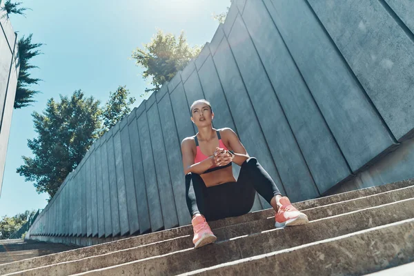 Vista de ángulo bajo de la hermosa mujer joven en ropa deportiva sentado al aire libre y mirando hacia otro lado —  Fotos de Stock