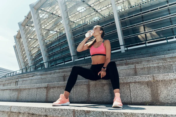 Beautiful young woman in sportswear drinking water while sitting outdoors — Stock Photo, Image