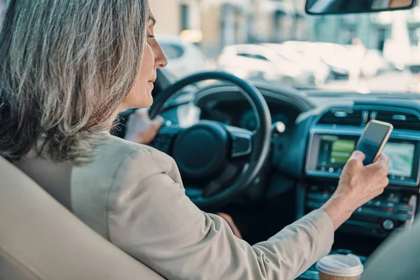 Pandangan belakang wanita cantik dewasa dengan pakaian kasual pintar menggunakan telepon saat mengendarai mobil — Stok Foto