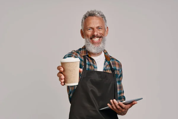 Senior man in apron smiling and giving you coffee while standing against gray background — Stock Photo, Image