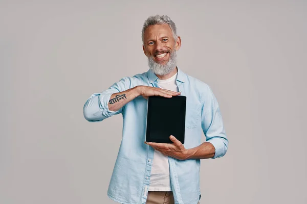 Happy senior man showing his digital tablet and smiling while standing against gray background — Stock Photo, Image