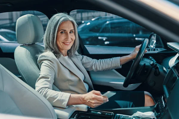 Starší krásná žena v chytré ležérní oblečení při pohledu na kameru a s úsměvem při řízení auta — Stock fotografie