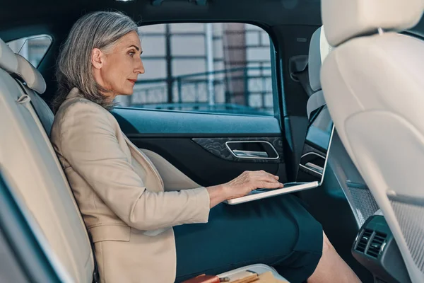 Confident mature woman in smart casual wear using laptop while sitting on the back car seat — 图库照片