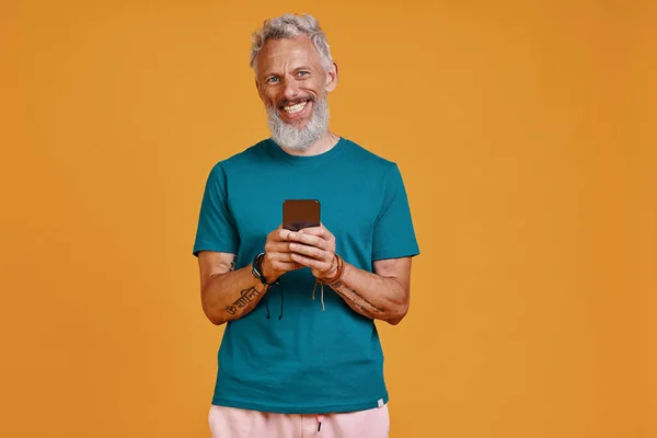 Felice uomo anziano utilizzando smart phone e sorridente mentre in piedi contro sfondo arancione — Foto Stock