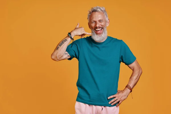 Feliz homem sênior fazendo me chame de gesto e sorrindo enquanto está de pé contra o fundo laranja — Fotografia de Stock