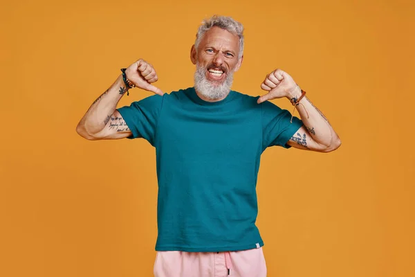 Happy senior man looking at camera and pointing himself while standing against orange background