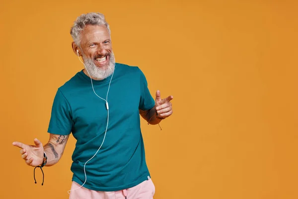 Happy senior man listening music and smiling while standing against orange background — Stock Photo, Image