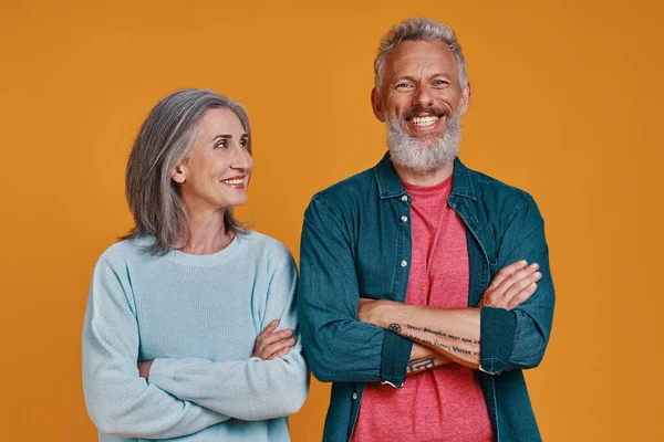 Beautiful senior couple keeping arms crossed and smiling while standing together against orange background — Stock Photo, Image
