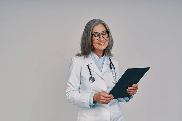 Madura hermosa doctora en bata de laboratorio blanca sonriendo mientras está de pie sobre fondo gris — Foto de Stock