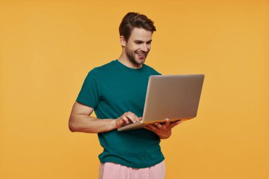Laptop kullanan ve sarı arka planda dururken gülümseyen sıradan giysili yakışıklı bir genç.