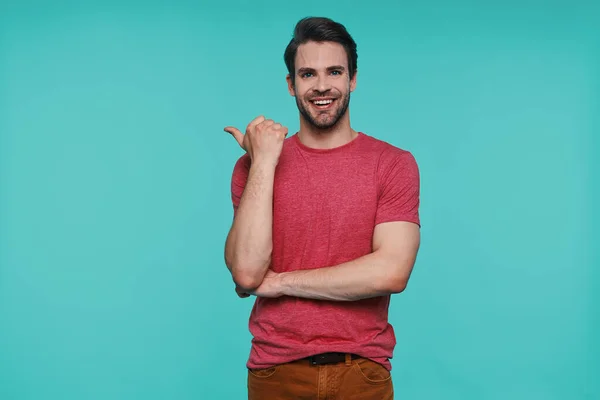 Handsome young smiling man in casual clothing looking at camera and pointing away — Stock Photo, Image