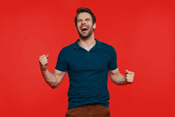 Happy young man in casual wear keeping eyes closed and gesturing while standing against red background — Stock Photo, Image